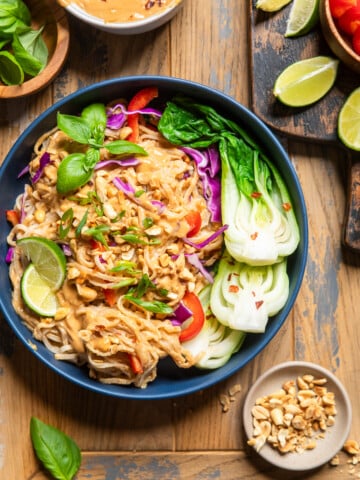vegan thai peanut noodles in a bowl on a table with ingredients surrounding to make.